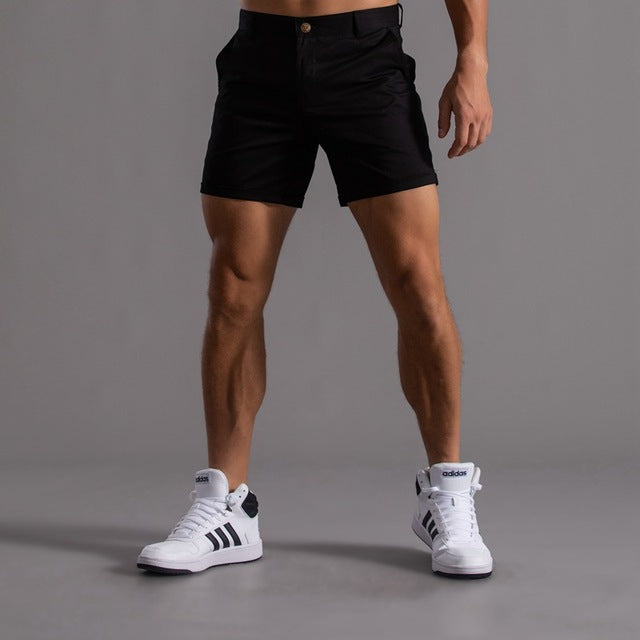Homme portant un short noir et des baskets blanches Adidas, debout sur un fond gris. Mode masculine, tenue décontractée, style sportif.