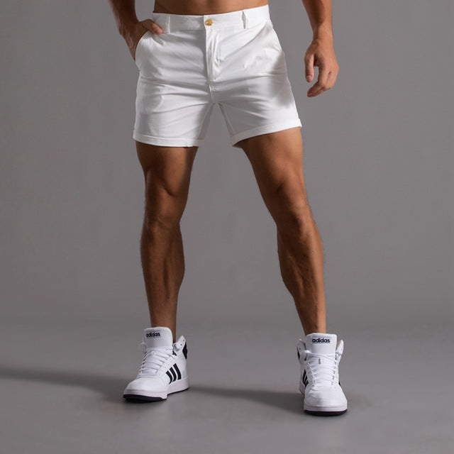 Homme portant un short blanc élégant et des baskets Adidas blanches, posant sur fond gris. Mode masculine, style décontracté, vêtements tendance.