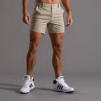 Homme portant un short beige et des baskets blanches Adidas sur fond gris. Mode masculine, tenue décontractée, style sportif.