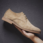 Chaussure derby en daim beige tenue en main, semelle marron clair, style élégant et moderne, idéale pour mode masculine et occasions formelles.