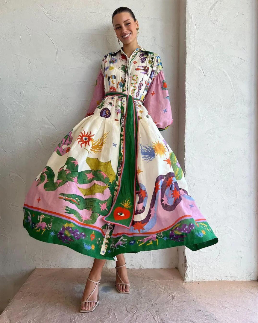 Robe longue colorée avec motifs floraux et animaliers, manches longues, ceinture verte, portée par une femme souriante, fond mural texturé.