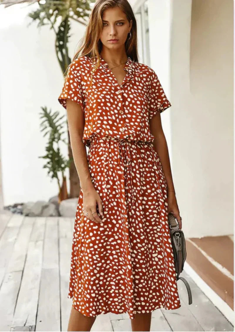 Femme portant une robe midi à motifs blancs sur fond marron, style décontracté, manches courtes, ceinture à la taille, posant sur une terrasse en bois.