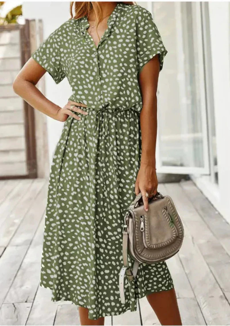 Robe verte à pois blancs, manches courtes, ceinture à la taille, portée par une femme tenant un sac à main beige sur une terrasse en bois.