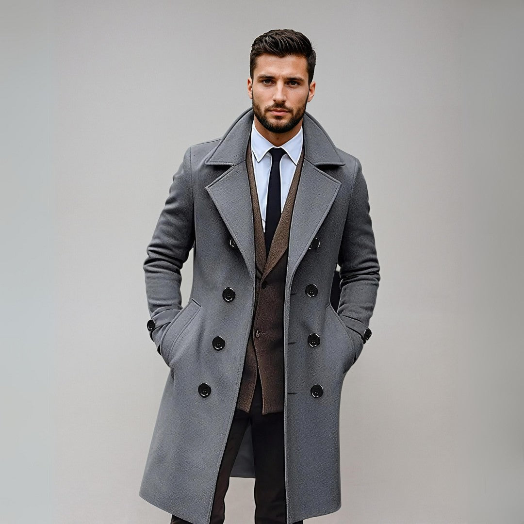 Homme élégant portant un manteau gris à double boutonnage, costume marron et cravate noire, posant sur fond neutre. Mode masculine chic.