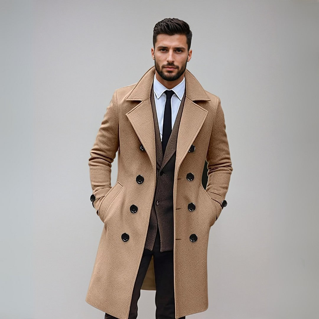 Homme élégant en manteau camel, costume marron et cravate noire, posant contre un mur gris. Mode masculine, style automne-hiver, tenue chic.