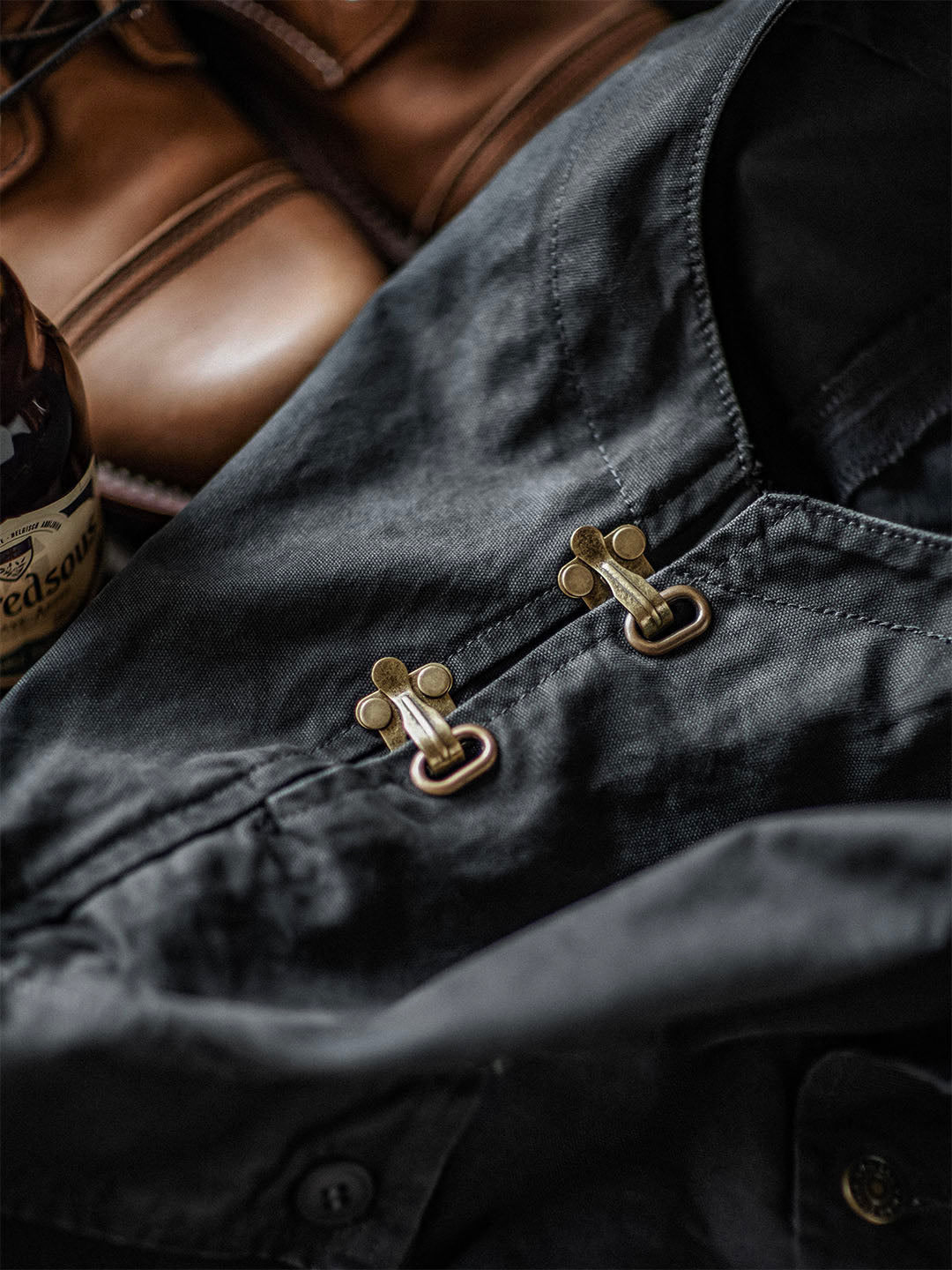 Chemise noire en toile avec boutons en métal doré, posée à côté de chaussures en cuir marron et d'une bouteille de whisky. Mode masculine élégante.