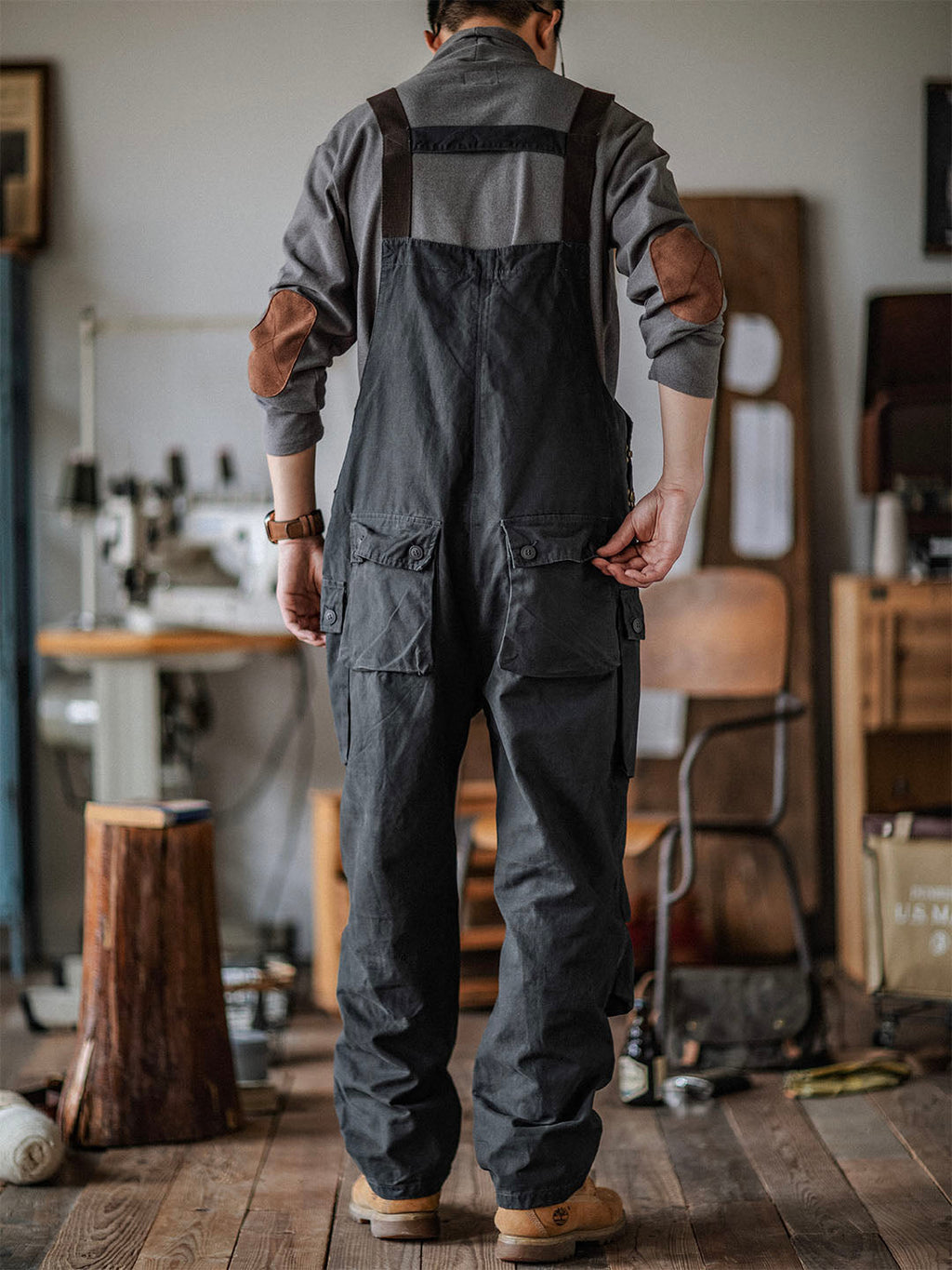 Homme en salopette noire dans un atelier de couture, debout sur un plancher en bois, avec machines à coudre et outils en arrière-plan.