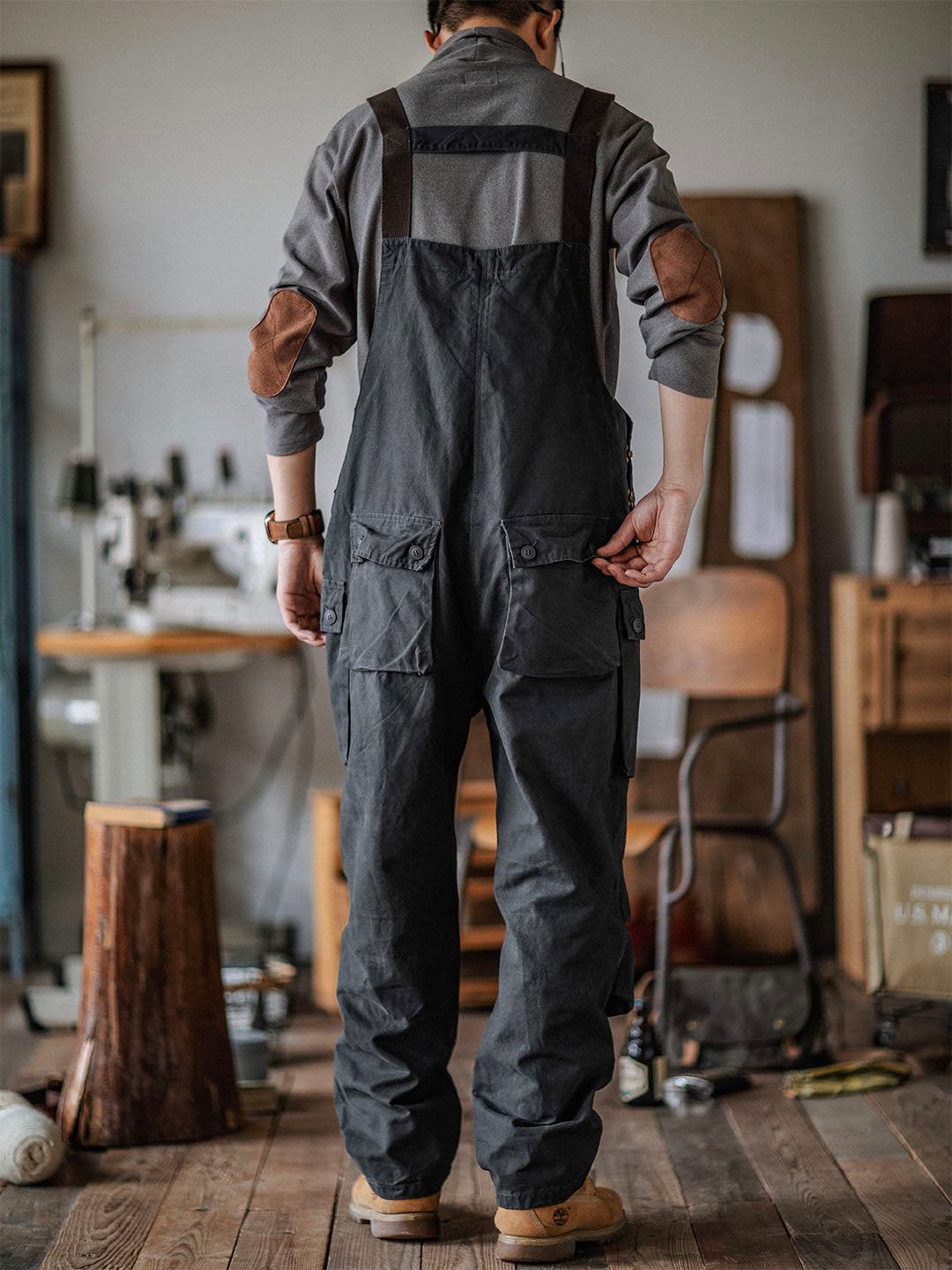 Homme en salopette noire dans un atelier de couture, debout sur un plancher en bois, avec machines à coudre et outils en arrière-plan.