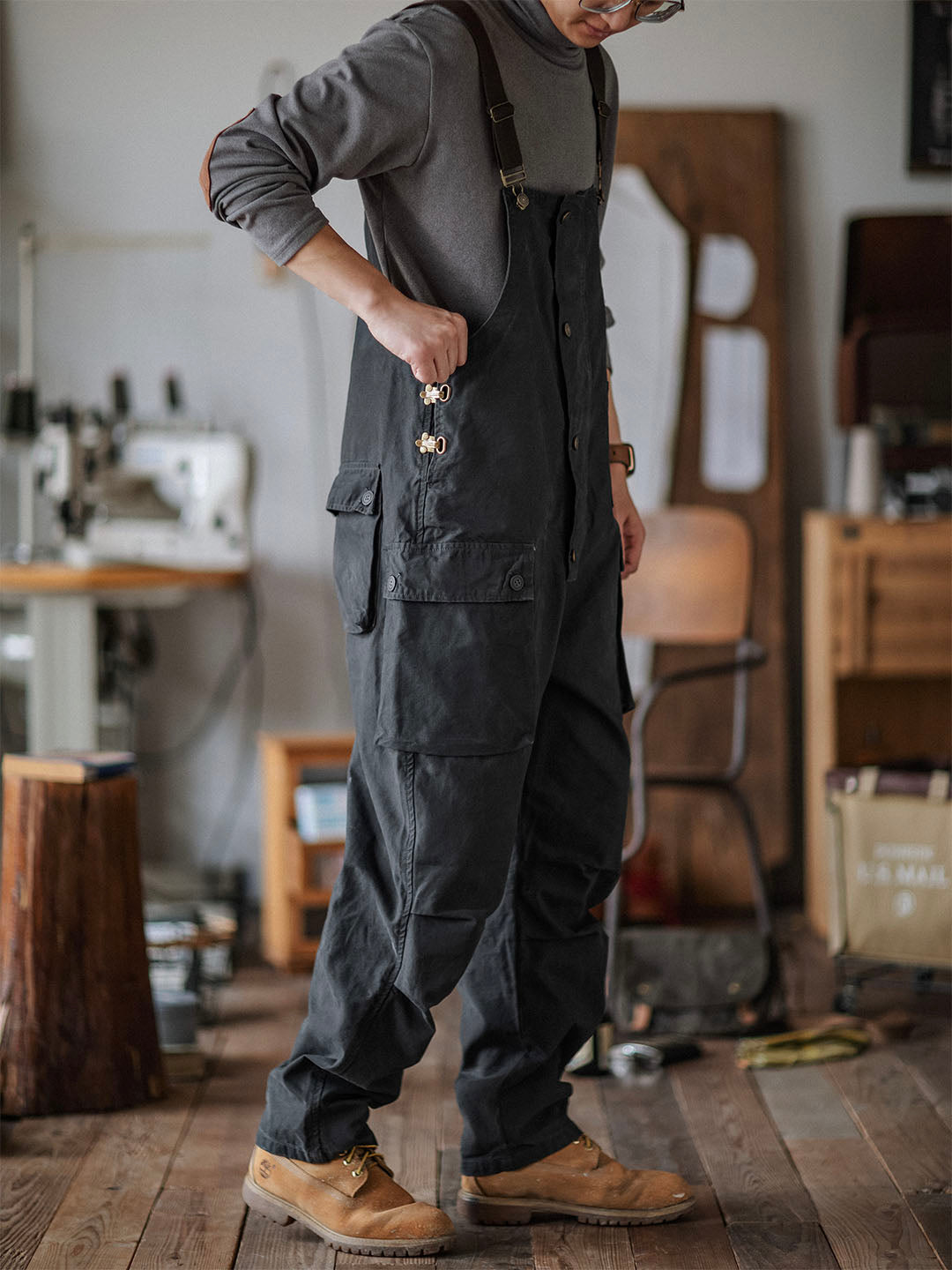Homme portant une salopette noire dans un atelier de couture, avec des bottes marron. Machine à coudre et outils en arrière-plan. Mode artisanale.