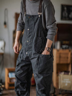 Homme portant une salopette noire avec poches, montre-bracelet visible, intérieur moderne en arrière-plan flou. Mode décontractée, style urbain.