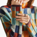 Femme portant un pull à carreaux colorés, main posée sur le bras, bague argentée visible. Mode automne-hiver, style décontracté et tendance.