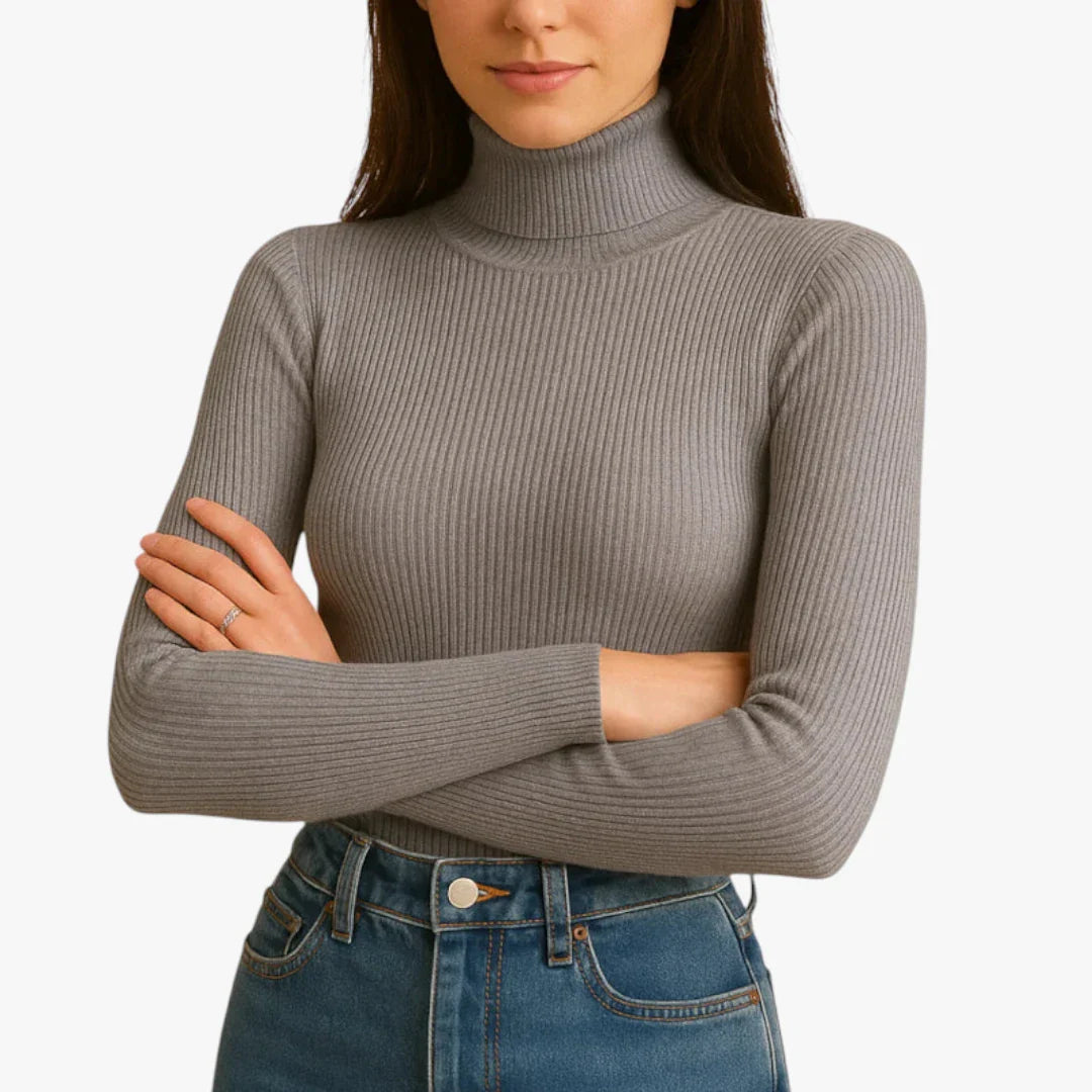Femme portant un pull col roulé gris côtelé, manches longues, bras croisés, associé à un jean bleu taille haute. Mode automne-hiver élégante.