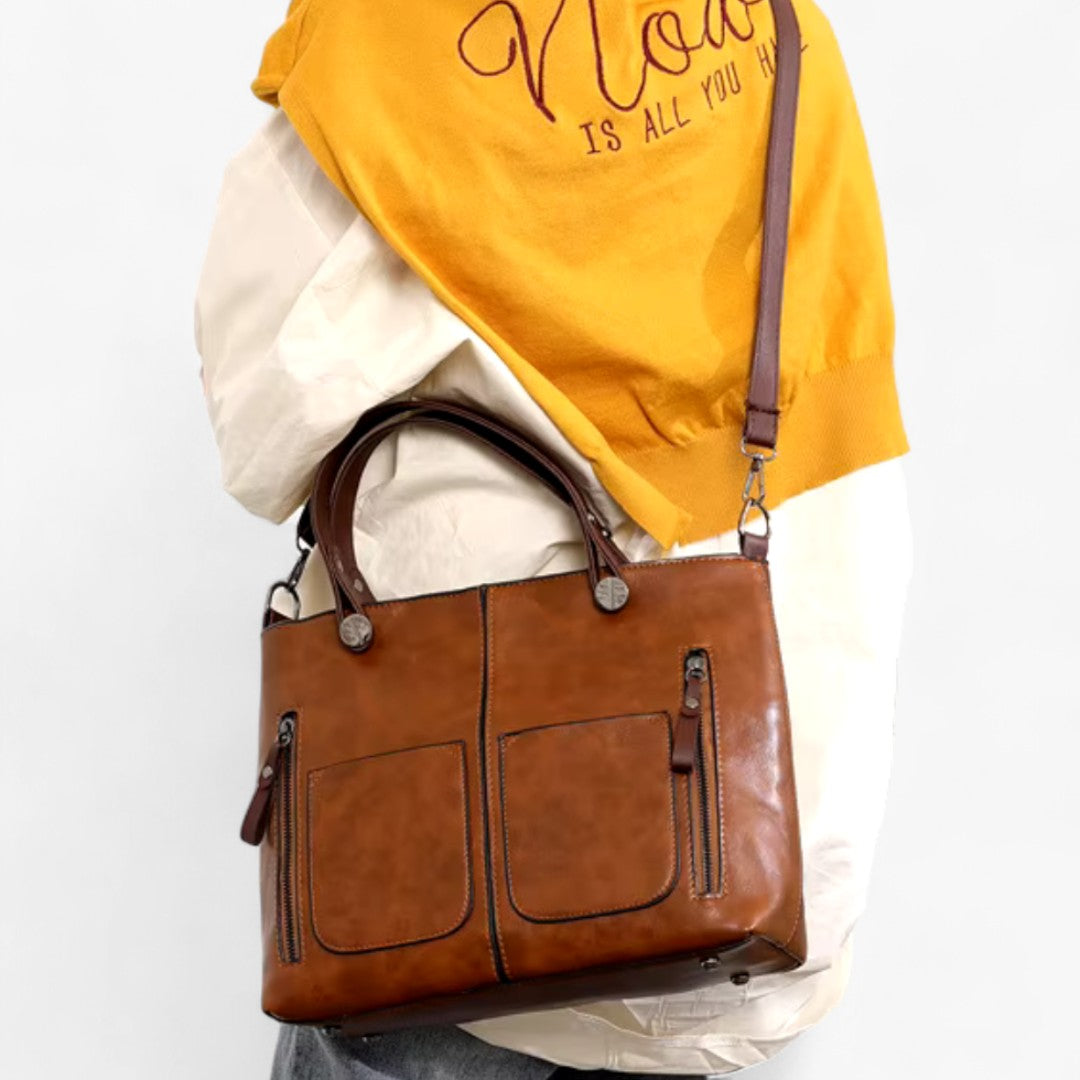 Sac à main en cuir marron avec deux poches avant et fermetures éclair, porté par une personne en pull jaune. Accessoire mode élégant et pratique.