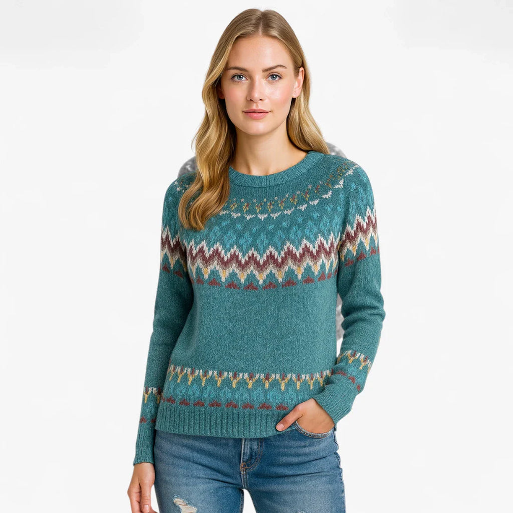 Femme portant un pull en laine bleu avec motifs géométriques colorés, manches longues, col rond, posant debout, jeans décontracté. Mode automne-hiver.