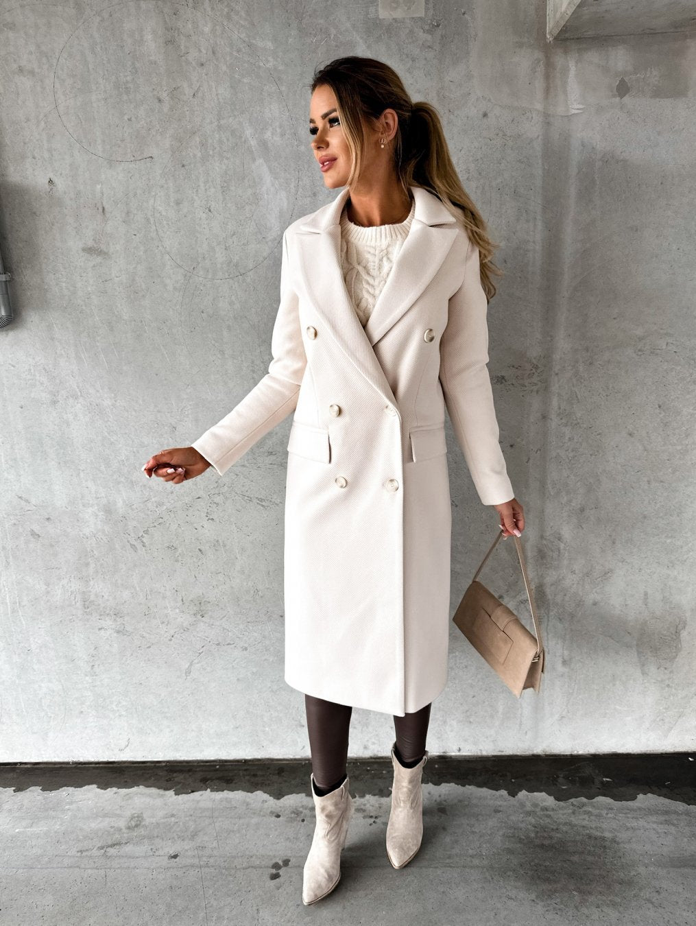 Femme portant un manteau blanc élégant, sac à main beige, bottines assorties, posant contre un mur gris. Mode automne-hiver, style chic et moderne.