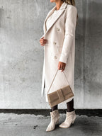 Femme portant un manteau long beige, tenant un sac à main assorti, avec des bottines élégantes, posant devant un mur gris texturé.