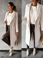 Femme élégante en manteau blanc, pull en maille crème, pantalon en cuir marron et bottines beiges, posant contre un mur gris. Mode automne chic.