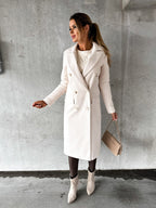 Femme portant un manteau blanc élégant, sac à main beige, bottines assorties, posant contre un mur gris. Mode automne-hiver, style chic et moderne.