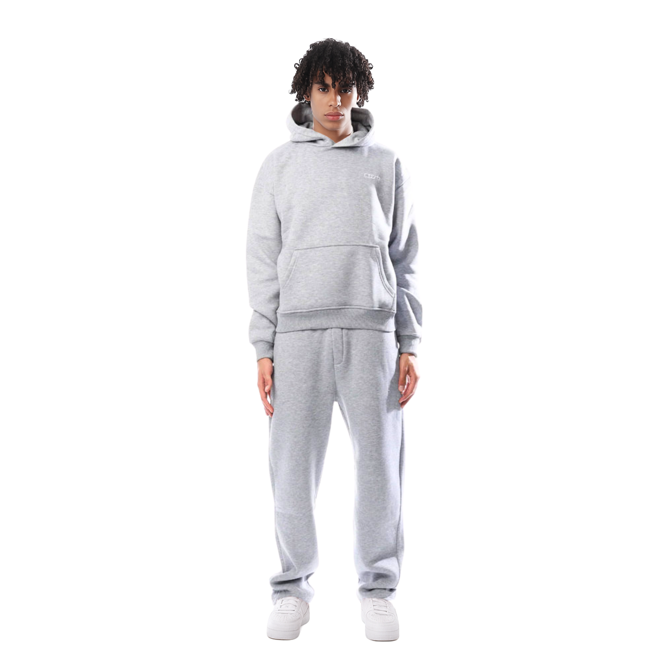 Jeune homme portant un survêtement gris, composé d'un sweat à capuche et d'un pantalon de jogging, debout sur fond blanc. Mode décontractée.
