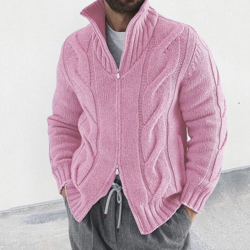 Homme portant un cardigan rose en tricot torsadé avec fermeture éclair, col montant, manches longues, sur un pantalon gris. Mode masculine élégante.