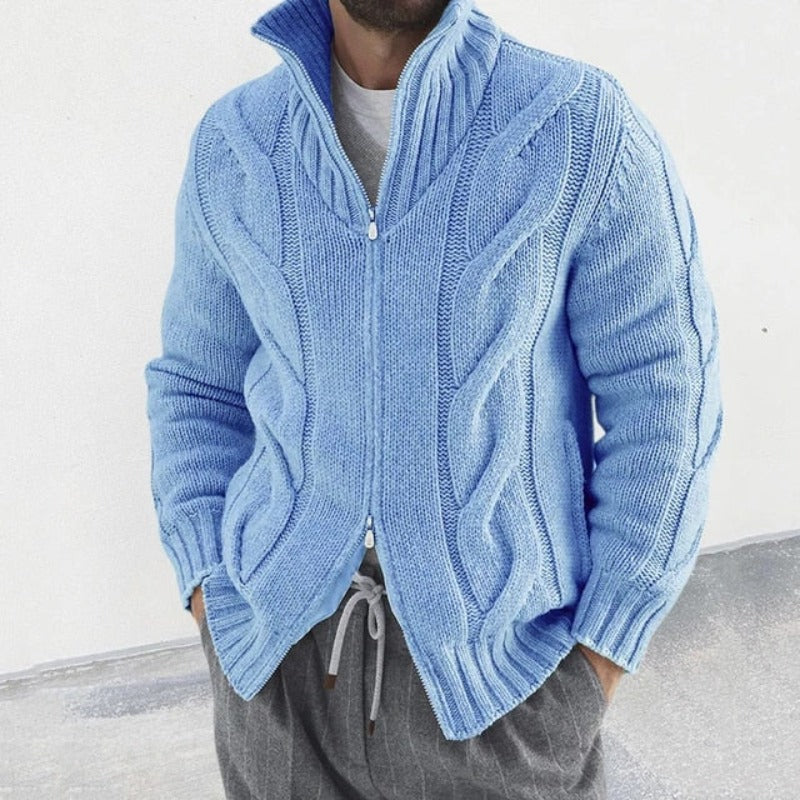 Homme portant un pull bleu en tricot torsadé avec fermeture éclair, col montant, manches longues, sur un t-shirt gris et pantalon gris.