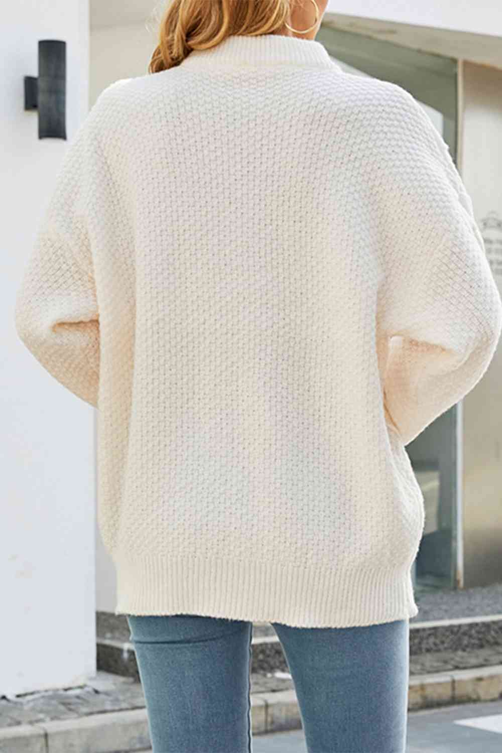Femme portant un pull blanc texturé, manches longues, style décontracté, vue de dos, avec un jean bleu, en extérieur urbain.
