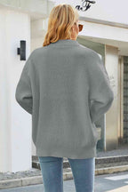 Femme de dos portant un pull gris texturé, manches longues, devant un bâtiment moderne. Mode automne, style décontracté, tendance féminine.