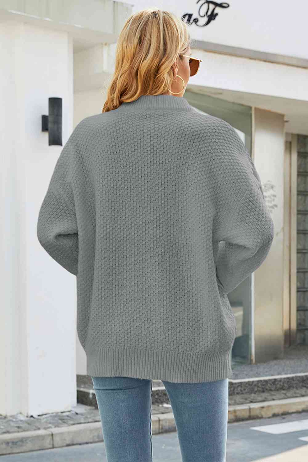 Femme de dos portant un pull gris texturé, manches longues, devant un bâtiment moderne. Mode automne, style décontracté, tendance féminine.
