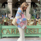 Femme portant une veste patchwork colorée et un sac beige, marchant devant un café parisien avec des chaises en osier. Mode urbaine chic.