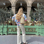 Femme de dos avec veste patchwork colorée et sac en paille, devant café parisien avec terrasse, chaises en osier, et architecture classique.