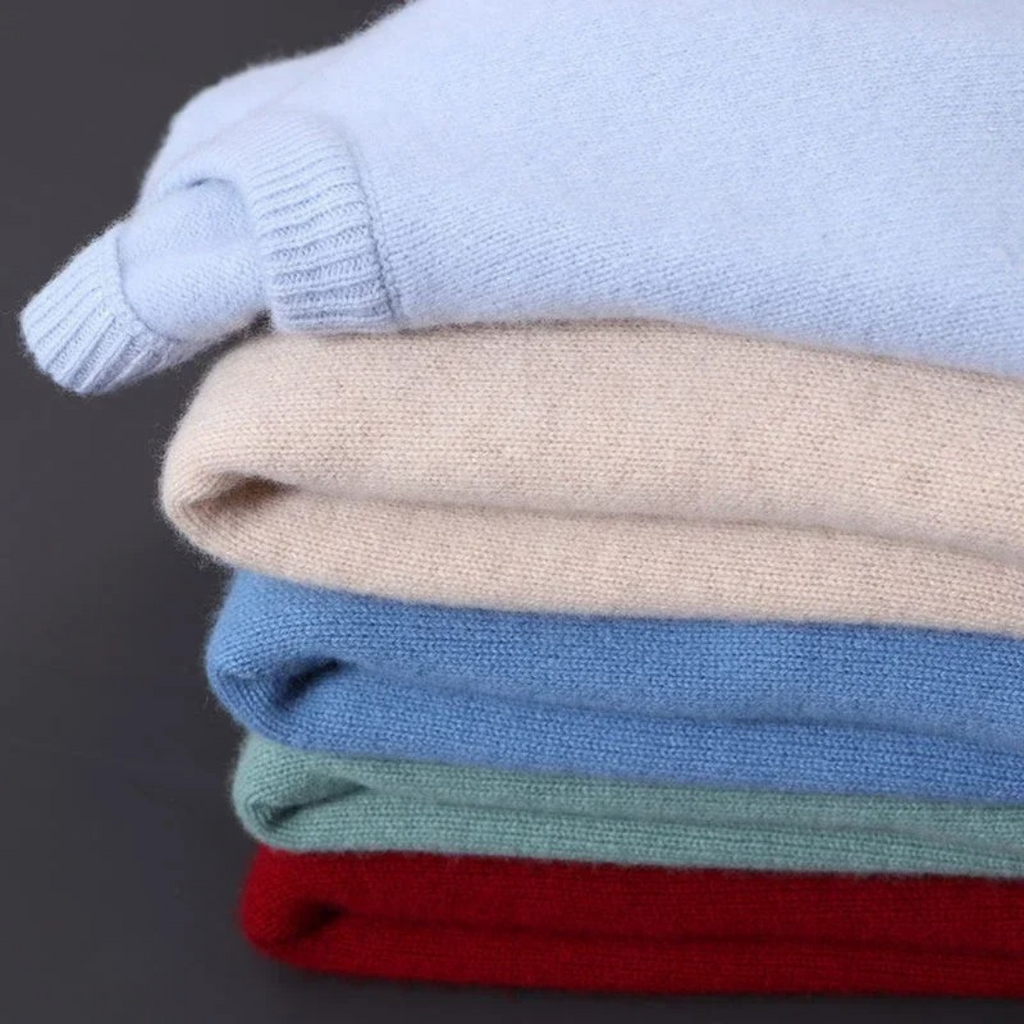Pile de pulls en cachemire colorés, doux et élégants, comprenant des teintes bleu clair, beige, bleu, vert et rouge, sur fond gris.