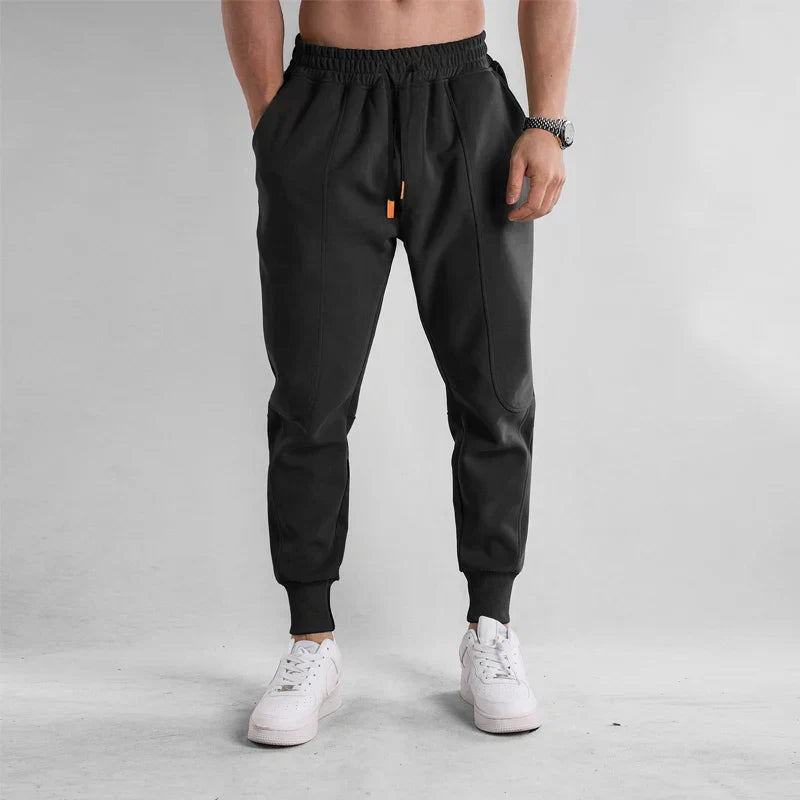 Homme portant un pantalon de jogging noir moderne avec baskets blanches, style décontracté, mode masculine, fond gris minimaliste.
