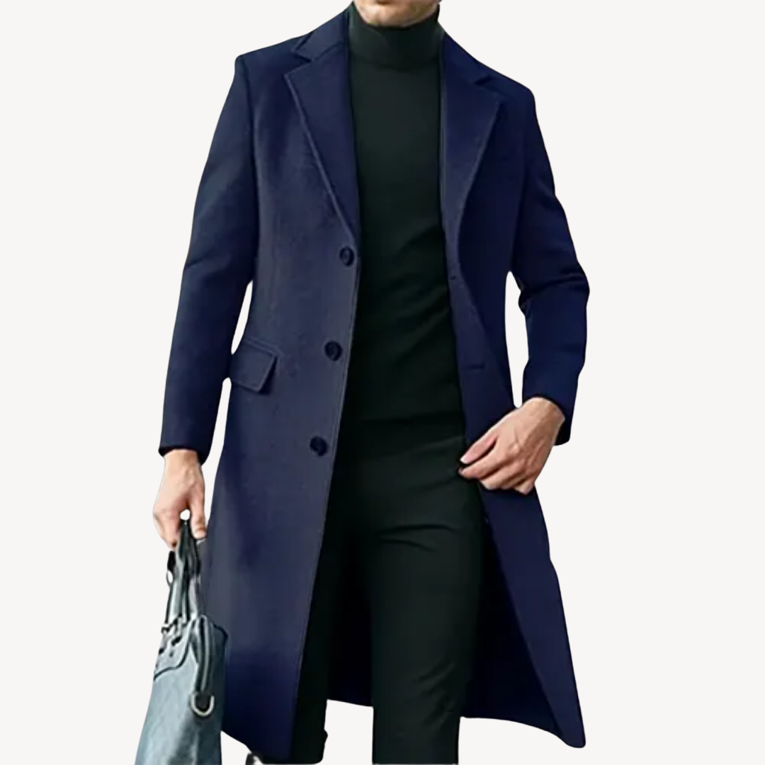 Homme portant un manteau long bleu marine élégant, avec col tailleur, sur un pull noir, tenant un sac gris. Mode masculine chic et moderne.