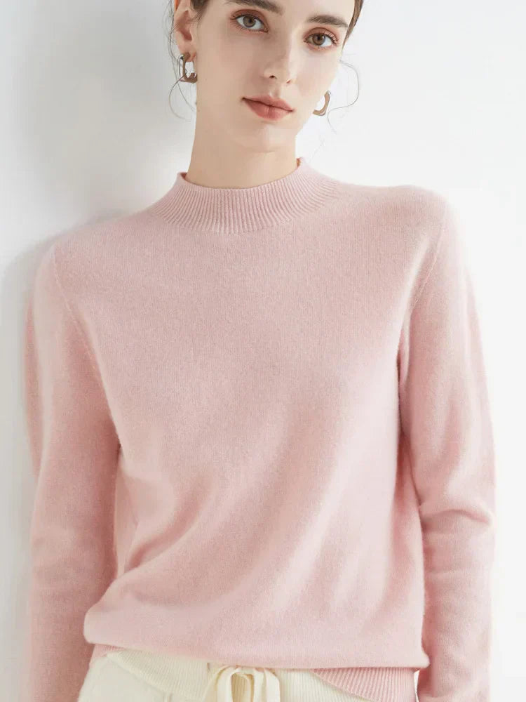 Femme portant un pull rose en cachemire, posant contre un mur blanc. Mode féminine élégante, tenue automne-hiver, style décontracté chic.
