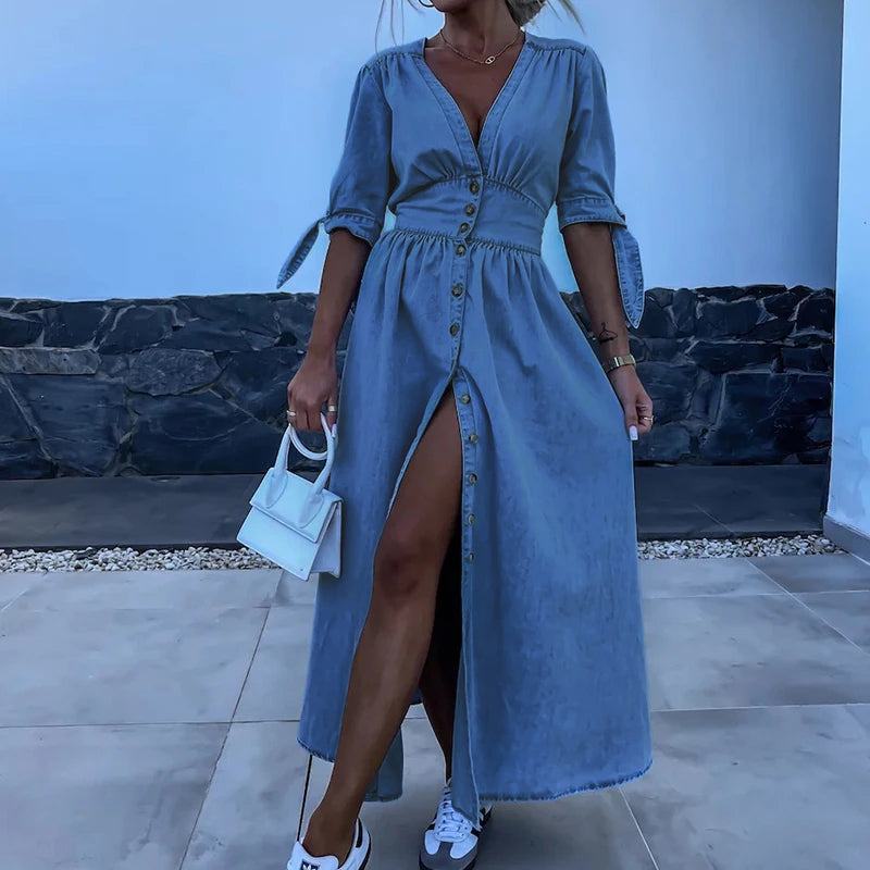 Robe longue en jean bleu avec boutons, fente avant, manches mi-longues, portée avec un sac blanc et des baskets, style décontracté chic.