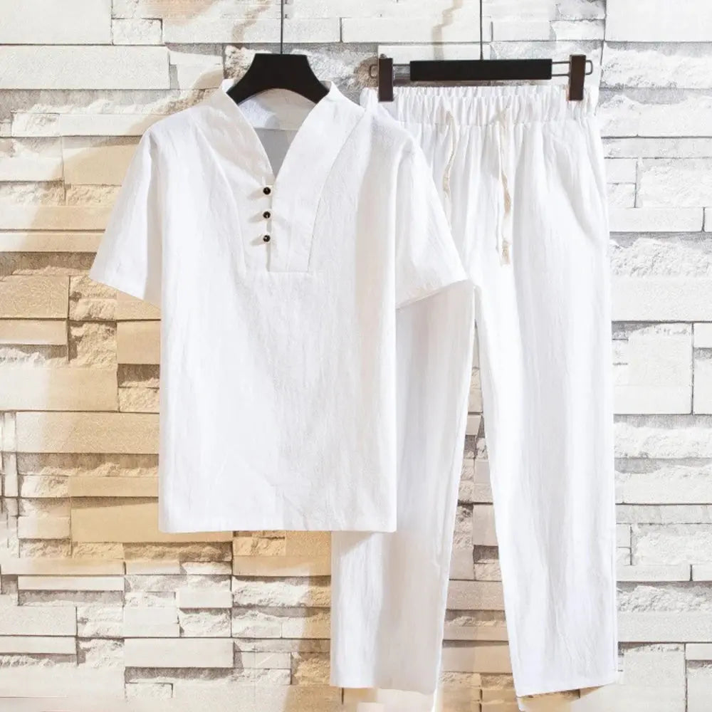 Tenue blanche en lin pour homme, chemise à col boutonné et pantalon assorti, suspendue sur un fond de mur en briques blanches.