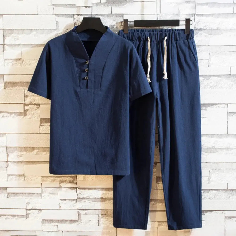 Tenue décontractée en lin bleu pour homme, comprenant une chemise à manches courtes avec col boutonné et un pantalon à cordon, suspendue sur un fond en briques blanches.