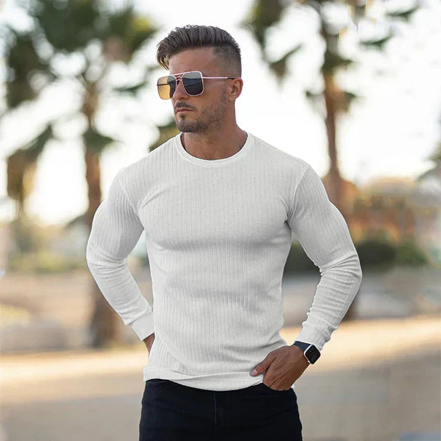 Homme portant un pull blanc ajusté, lunettes de soleil et montre noire, debout à l'extérieur avec des palmiers en arrière-plan. Mode masculine décontractée.
