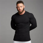 Homme musclé portant un pull noir ajusté, manches longues, sur fond gris. Mode masculine, vêtements fitness, style décontracté.
