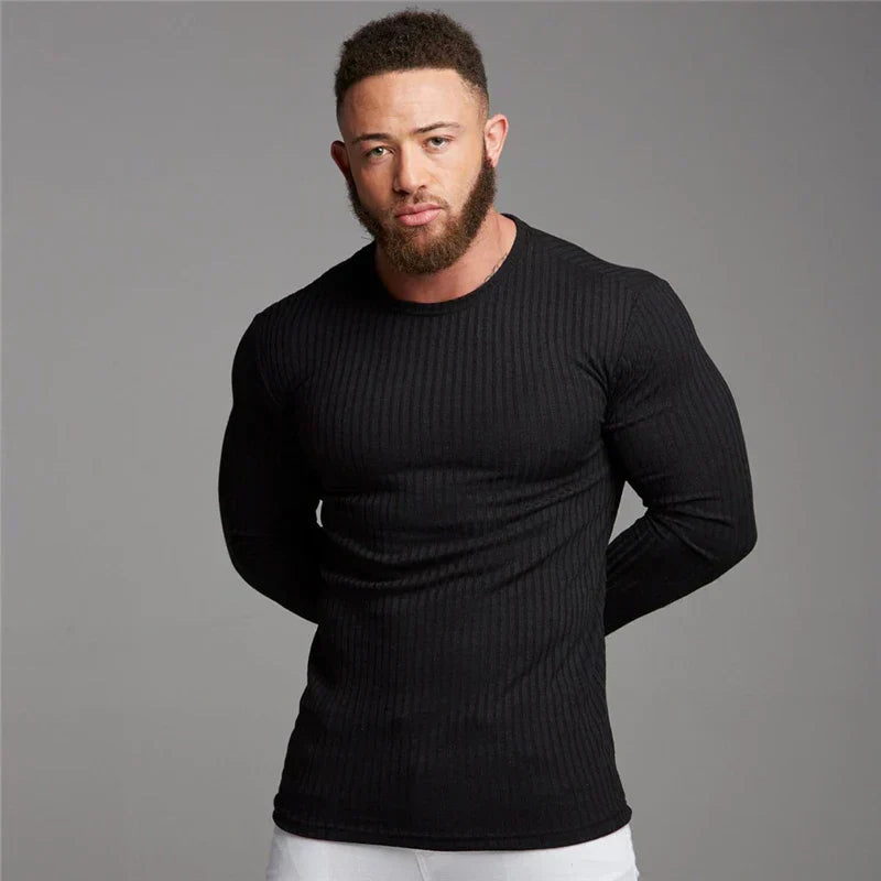 Homme musclé portant un pull noir ajusté, manches longues, sur fond gris. Mode masculine, vêtements fitness, style décontracté.