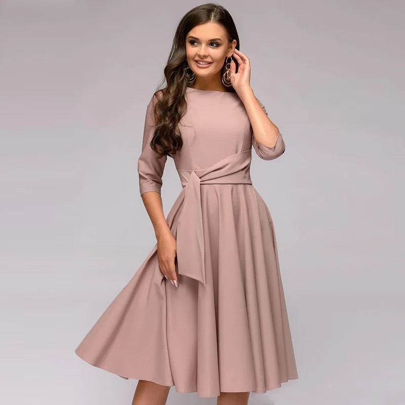 Robe élégante rose poudré, manches trois-quarts, ceinture nouée, portée par une femme souriante. Mode féminine chic, tendance automne.