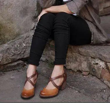 Femme assise portant des chaussures à talons marron croisés, pantalon noir, veste grise, assise sur un escalier en pierre. Mode automne chic.