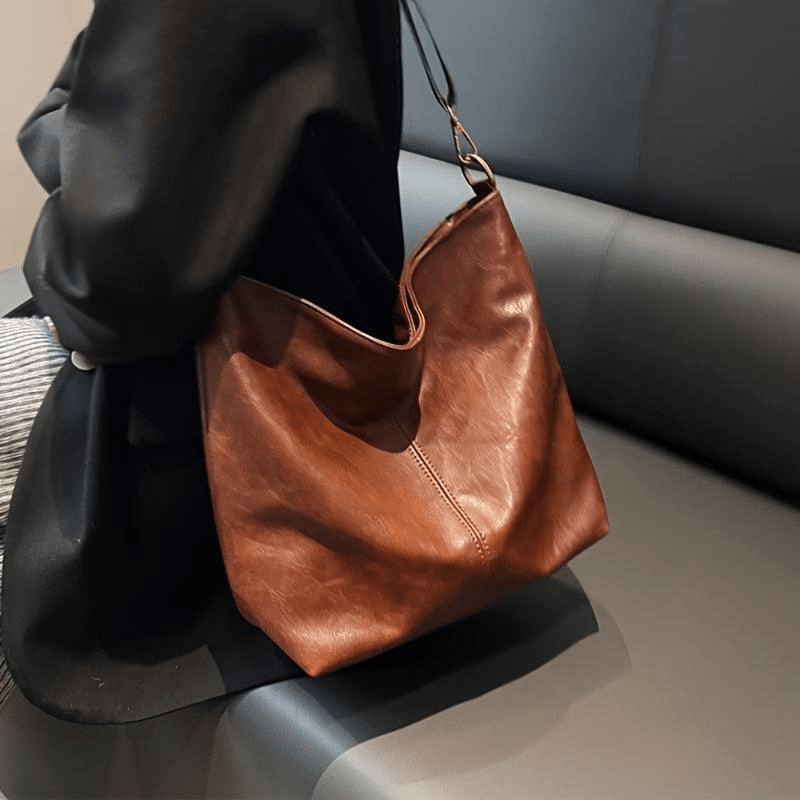 Sac à main en cuir marron porté par une personne assise sur un canapé noir. Accessoire de mode élégant, tendance et pratique pour le quotidien.