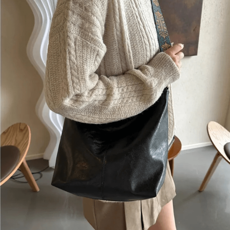 Femme portant un sac à main en cuir noir, style décontracté, avec un pull beige et une jupe marron. Mode automne, accessoires tendance.