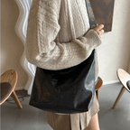 Femme portant un sac à main en cuir noir, style décontracté, avec un pull beige et une jupe marron. Mode automne, accessoires tendance.