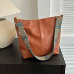 Sac à main en cuir marron avec bandoulière brodée bleue et dorée, posé sur une table noire près d'une lampe beige. Accessoire mode élégant.