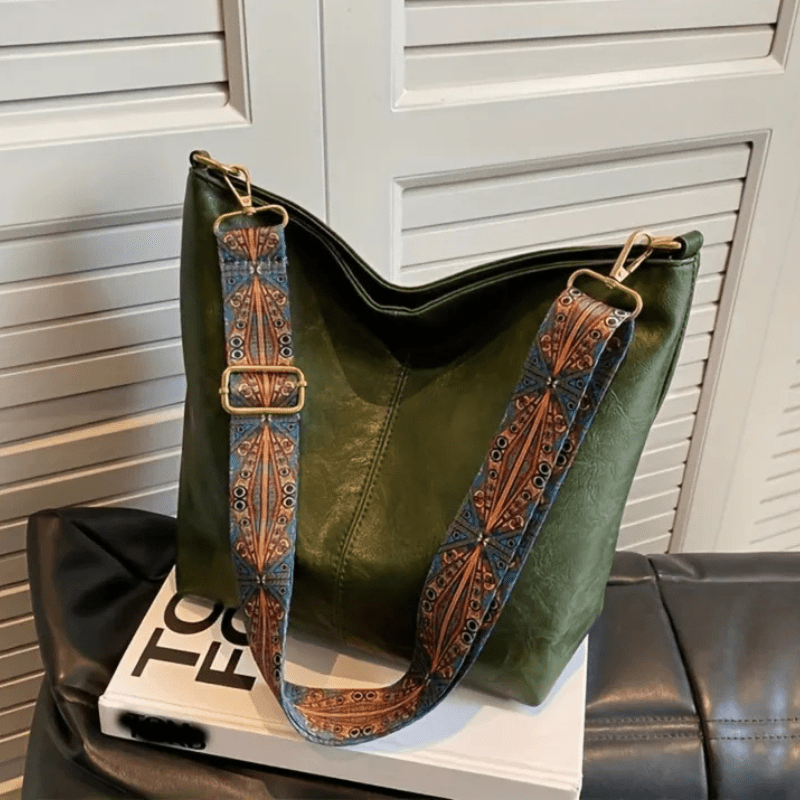 Sac à main vert en cuir avec bandoulière brodée, posé sur un livre blanc. Accessoire de mode élégant, tendance et chic pour femmes.