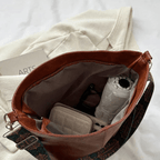 Sac à main en cuir marron ouvert, contenant un livre, un parapluie blanc, un portefeuille beige et des lunettes de soleil, posé sur un tissu blanc.