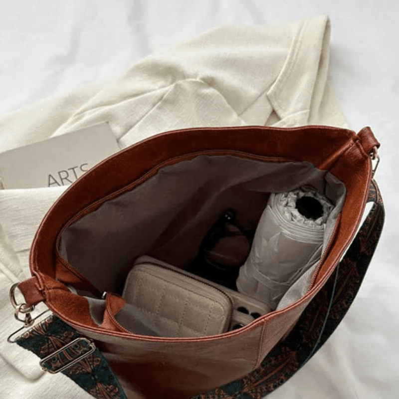 Sac à main en cuir marron ouvert, contenant un livre, un parapluie blanc, un portefeuille beige et des lunettes de soleil, posé sur un tissu blanc.