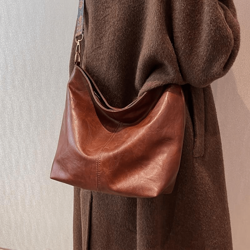 Sac à main en cuir marron porté sur l'épaule d'une personne vêtue d'un manteau en laine marron. Accessoire mode élégant et tendance.